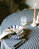 A styled dining table setting with a blue and white gingham tablecloth, a matching blue and white striped ceramic plate with a beige cloth napkin, black cutlery, a small clear glass, and two lit white candlesticks.