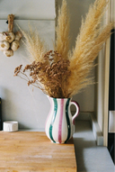 A white ceramic pitcher with vertical pink, green, and black stripes, filled with dried pampas grass and brown botanicals, set on a wooden cutting board on a kitchen counter next to a bottle of olive oil.