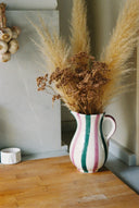 A white ceramic pitcher vase with vertical pink, green, and dark stripes, filled with dried pampas grass and brown botanicals, set on a wooden tabletop against a gray wall.