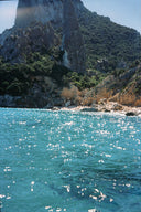 A scenic coastal photograph showing bright turquoise water in the foreground and a steep rocky cliff covered with green vegetation rising behind it under a pale blue sky.