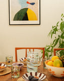 A framed abstract art print hangs on a beige wall above a wooden dining table set with bowls, glasses, and a fruit bowl, with green plants around the table.