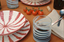 A styled dining table setting featuring a set of white ceramic plates and glasses with red radial stripes, alongside a dark wine bottle, a small vine of cherry tomatoes, and a folded beige napkin on a brown tablecloth.
