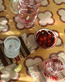 A styled tabletop scene with a glass pitcher of pink drink, a bowl of red cherries, two glasses with ice, and a lime wedge on a patterned yellow tablecloth.