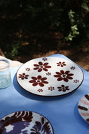 A white ceramic plate with a dark brown floral pattern, set on a blue tablecloth outdoors with a glass cup nearby.