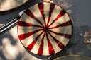 A round glass plate with a cream-colored base and a red radial starburst pattern, styled on a gray table with silver cutlery and other dining items.