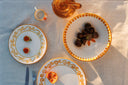 Three white ceramic plates with gold scrollwork borders are arranged on a light beige linen tablecloth, styled with a small orange cup, a glass teapot, and food items under warm sunlight.
