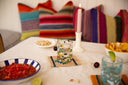 A styled tabletop scene with colorful striped pillows in the background, a lit pink taper candle in a white holder, a glass tumbler with multicolored stones, bowls of salsa and chips, and a blue-tinted drink in the foreground on a white tablecloth.