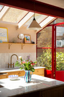 A bright kitchen interior with a marble countertop in the foreground, a blue-and-white striped vase holding colorful flowers, wooden cabinetry, a sink, and a red-framed glass door opening to greenery outside.